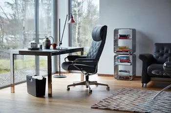 Stressless Metro Office-Stuhl in der Farbe Paloma Metal Grey mit verchromtem Gestell auf Rollen. Der Stuhl steht an einem modernen Schreibtisch mit großen Glasfenstern im Hintergrund. Bücherregal, Sofa und Tischlampe ergänzen die stilvolle Einrichtung.